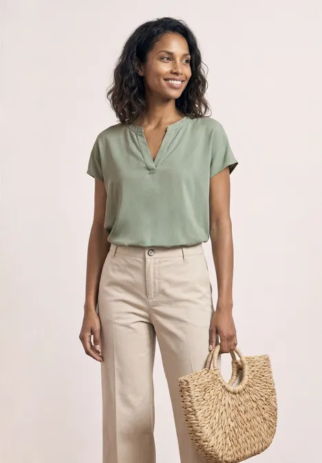 Eine Frau steht in leichter, sommerlicher Kleidung mit grüner Bluse und beigen Hosen und hält eine geflochtene Tasche.