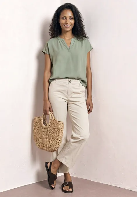 Frau lehnt an hellem Hintergrund, trägt grüne Bluse, helle Hose, schwarze Sandalen und einen geflochtenen Korb.