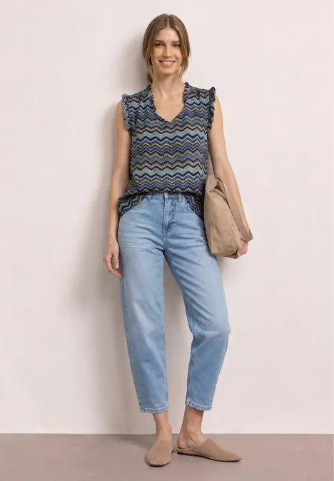 Eine Frau mit geblümtem Top, hoch taillierten Jeans, beigen Schuhen und Tasche vor neutralem Hintergrund.