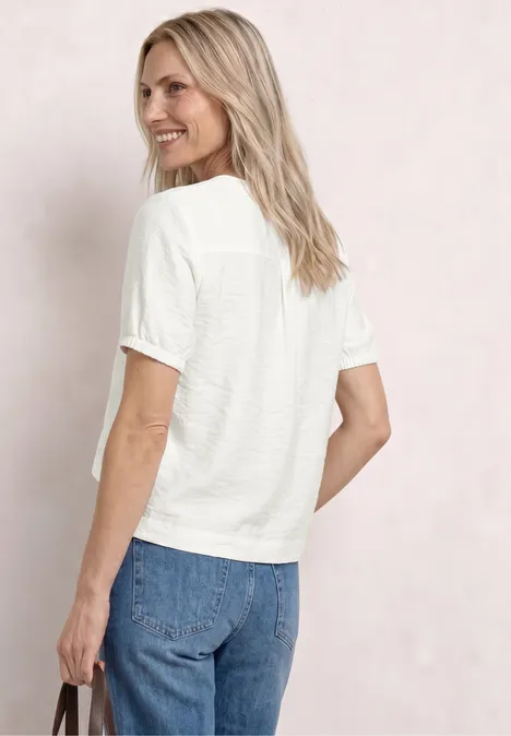 Frau von hinten in weißem T-Shirt und blauen Jeans, hält schwarze Tasche, lockere blonde Haare, neutraler Hintergrund.