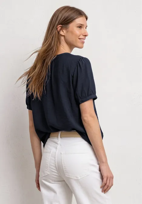 Frau von hinten mit dunkelblauem T-Shirt und weißer High-Waist-Hose vor neutralem Hintergrund.
