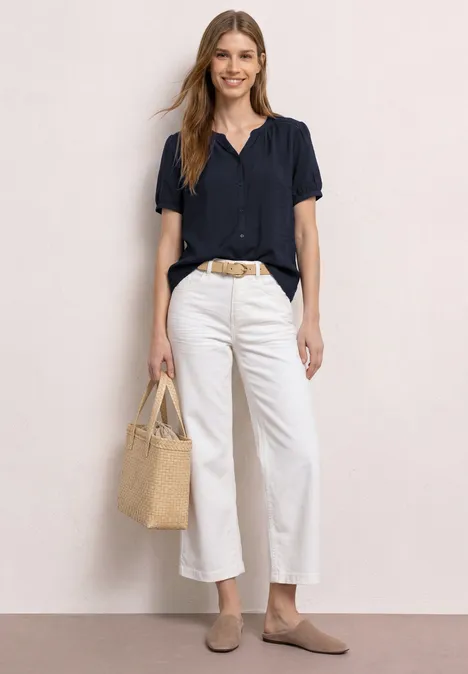 Frau in dunkelblauer Bluse, weißen Culottes, beigen Loafers und Basttasche vor hellem Hintergrund.
