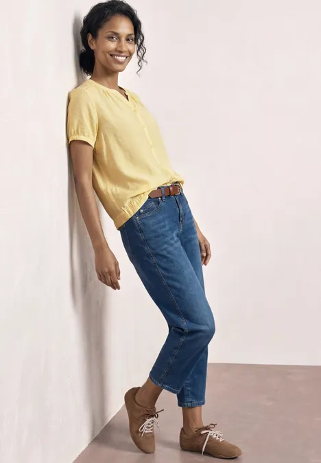 Frau lehnt entspannt in gelbem Shirt, blauen Jeans und braunen Schuhen an heller Wand.