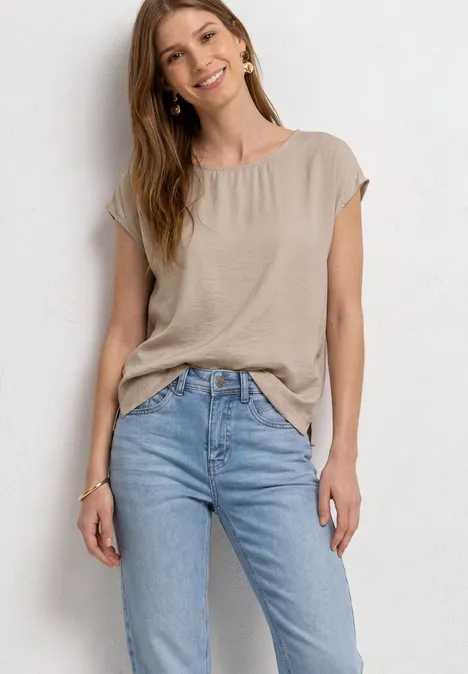 Eine Frau trägt eine beige Kurzarmbluse und hellblaue Jeans und steht entspannt vor hellem Hintergrund.