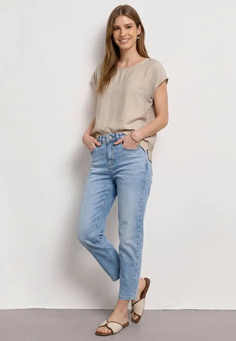 Frau in beigem Shirt und hellen Jeans, mit Sandalen, steht locker vor weißer Wand, Hände in den Taschen.