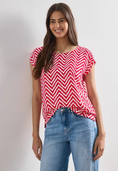 Eine Frau trägt ein rot-weiß gestreiftes Zickzack-Shirt und blaue Jeans vor hellem Hintergrund.