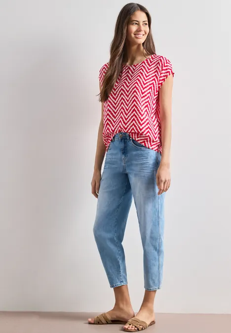 Eine Frau trägt ein rotes, weißes Zickzack-T-Shirt, blaue Jeans und Sandalen vor weißem Hintergrund.