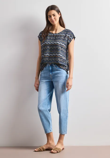 Frau steht vor grauem Hintergrund, trägt gemustertes Oberteil, blaue gekrempelte Jeans und Sandalen.