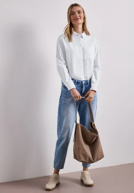 Frau trägt weiße Bluse, weite blaue Jeans, beige Schuhe und hält eine braune Tasche.