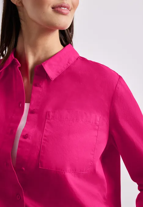 Vrouw draagt een felroze overhemd met kraag, knopen en zak, eronder een wit topje.
