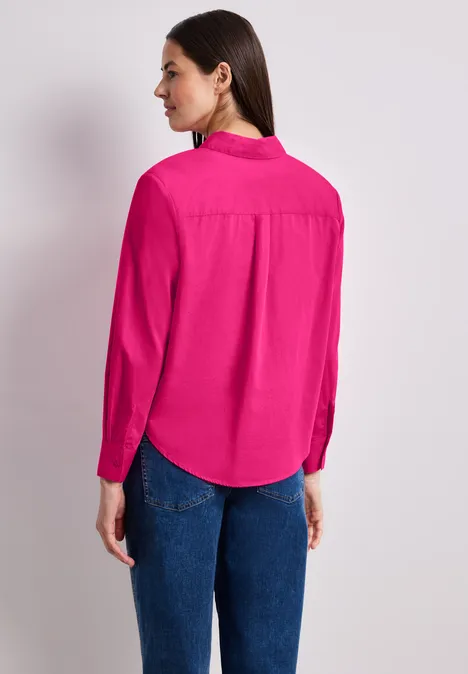 Een vrouw van achteren gefotografeerd draagt een felroze blouse en blauwe jeans voor een neutrale achtergrond.
