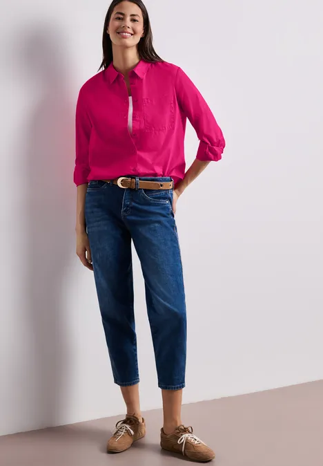 Vrouw in roze blouse, donkerblauwe spijkerbroek en bruine loafers voor een lichte achtergrond.