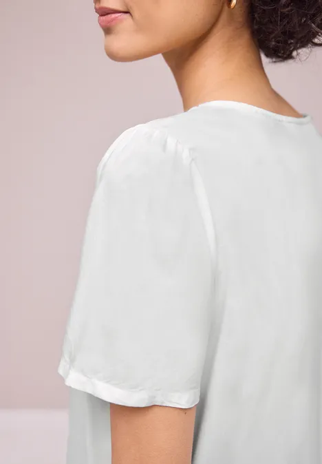 Een vrouw met krullend haar draagt een witte, soepel vallende korte mouwblouse, van opzij en van achter gefotografeerd.