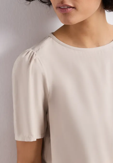 Vrouw draagt crèmekleurige blouse met ronde hals en korte mouwen, neutrale achtergrond.