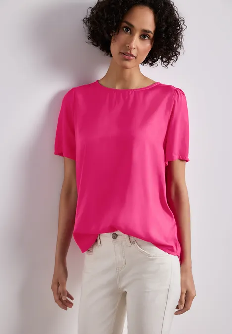 Persoon met krullend haar draagt een roze t-shirt en een witte broek voor een neutrale achtergrond.