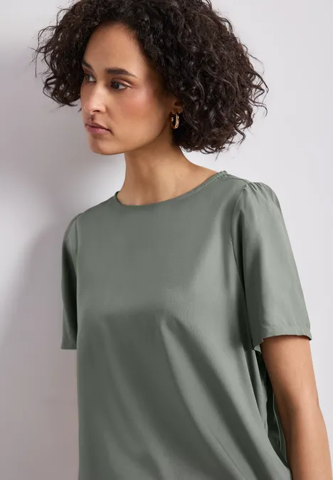 Een vrouw met krullend haar draagt een eenvoudig kort olijfgroen T-shirt voor een lichte achtergrond.