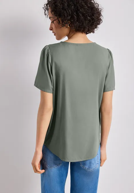 Vrouw van achteren in een loszittend grijsgroen T-shirt met korte mouwen en een blauwe spijkerbroek.