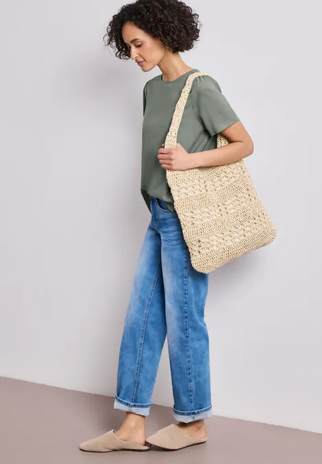 Persoon met groen T-shirt, blauwe jeans en beige instappers draagt grote gevlochten strozak.