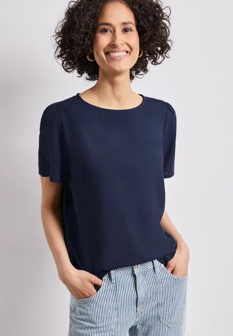 Frau steht mit Händen in den Hosentaschen, trägt dunkelblaue Kurzarm-Bluse und helle Jeans.