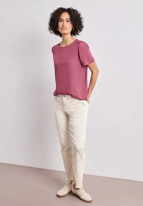 Bluse mit Rundhals und kurzen Puffärmeln dark clouded blush Frau steht vor neutralem Hintergrund, trägt rosa T-Shirt, weiße Hose und weiße Sneaker.