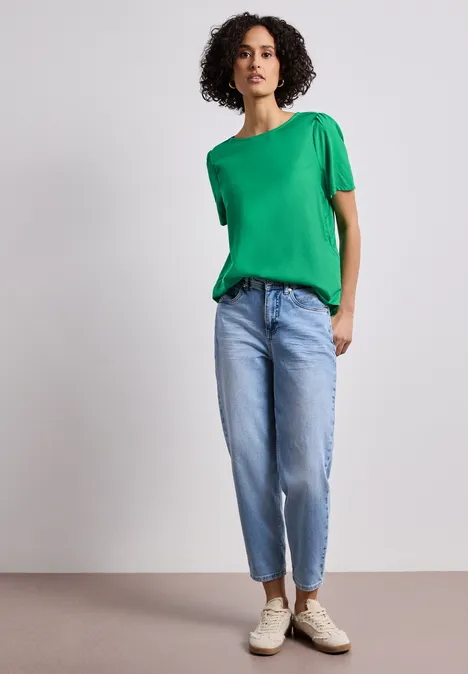 Bluse mit Rundhals und kurzen Puffärmeln ever green Eine Person in grünem Shirt, hellen Jeans und Sneakern steht vor neutralem Hintergrund.