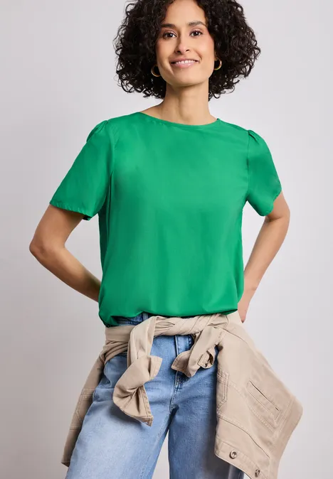 Bluse mit Rundhals und kurzen Puffärmeln ever green Person mit lockigen Haaren trägt grünes T-Shirt, Jeans und beige Jacke um die Taille, vor neutralem Hintergrund.
