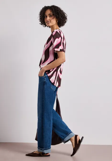 Persoon draagt een abstracte blouse met zijsplit, wijde jeans en zwarte sandalen, zijaanzicht.