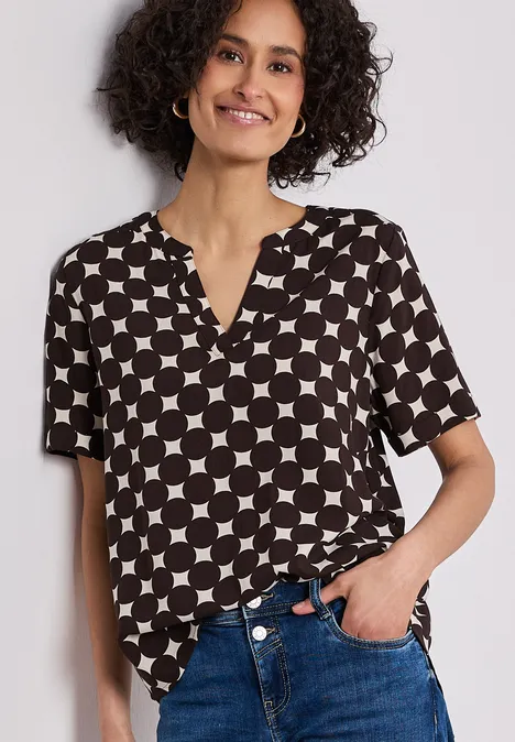 Vrouw draagt een zwart-witte blouse met korte mouwen en geometrisch cirkel- en vierkantmotief, deels in jeans gestopt.