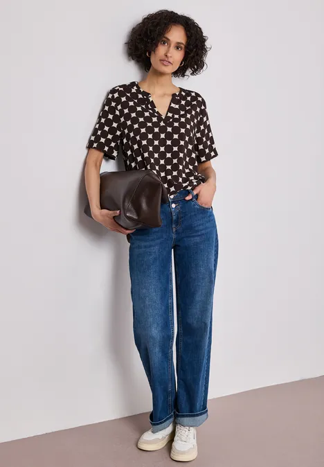 Vrouw met krullend haar, zwart blouse met witte harten, spijkerbroek, witte sneakers en tas leunend tegen muur.