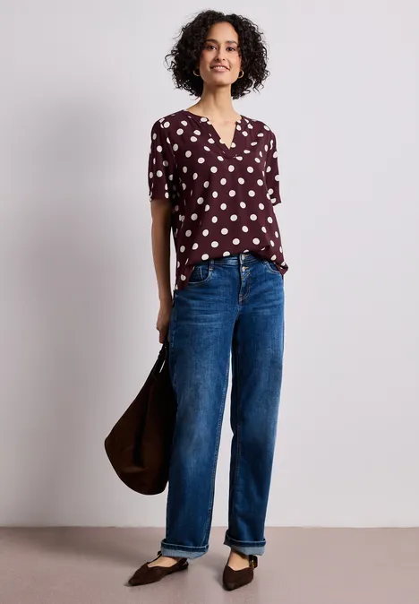 Vrouw in bordeaux blouse met witte stippen, blauwe jeans, zwarte ballerina’s en bruine tas.