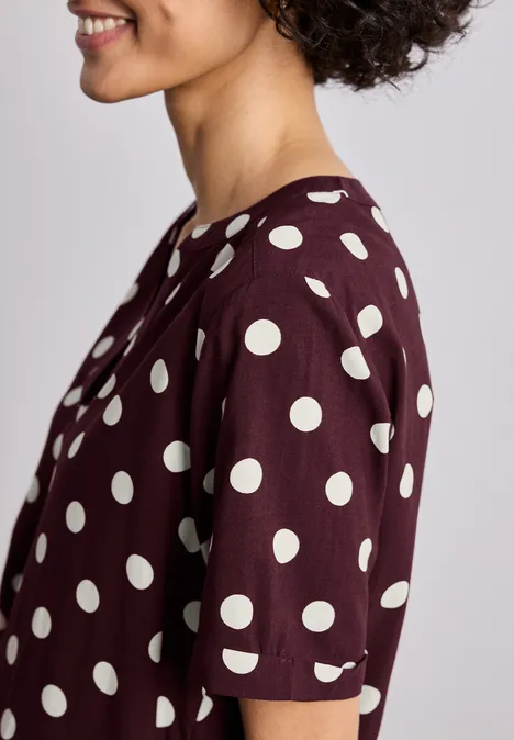 Zijdelings portret van een glimlachende persoon in een bordeaux t-shirt met witte stippen.