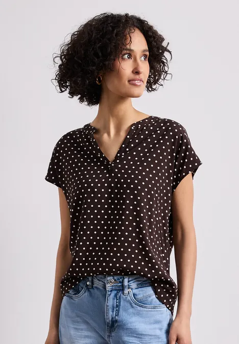 Vrouw met krullend haar draagt een polkadot blouse en blauwe spijkerbroek voor een neutrale achtergrond.