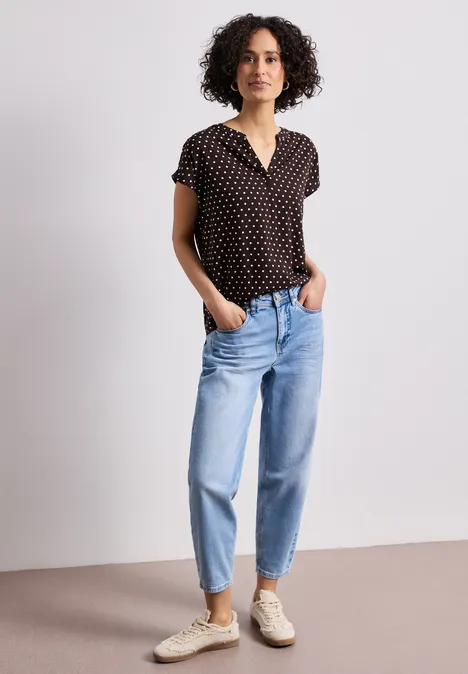 Een vrouw met krullend halflang haar draagt een zwart gestipt shirt, blauwe jeans en witte sneakers.