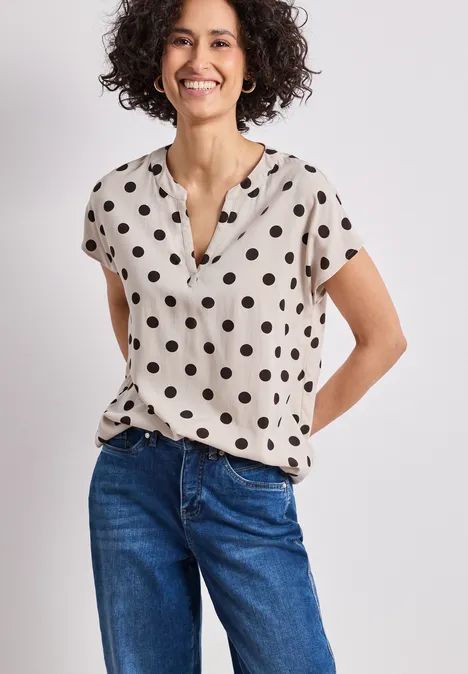 Vrouw in blauwe jeans en beige blouse met zwarte stippen en korte mouwen, handen achter de rug.