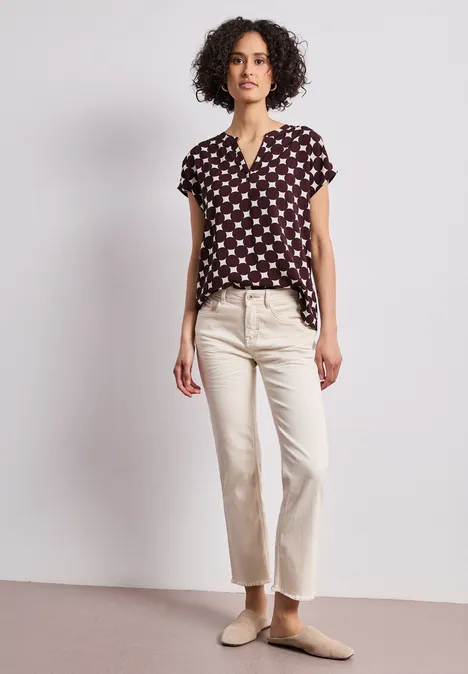 Dropped Shoulder Bluse mit Split Neck rustic rouge Eine Frau mit dunklem, lockigem Haar trägt eine schwarze Bluse mit weißem Muster, helle Hose und Pantoffeln.