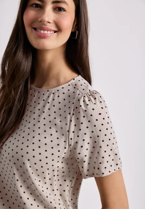 Frau trägt helles kurzärmeliges Oberteil mit schwarzen Polka-Dots, rundem Ausschnitt und leicht gerafften Schultern.