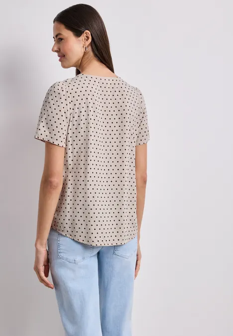 Frau von hinten in kurzer, gepunkteter Bluse und hellblauer Jeans vor neutralem Hintergrund.