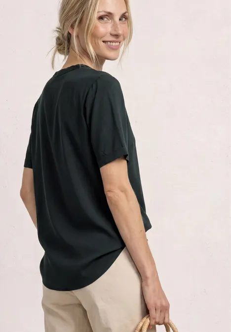 Vrouw in los zwart T-shirt en beige broek, licht schuin, voor lichte achtergrond.