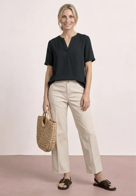 Vrouw in zwarte top, beige broek, zwarte sandalen en rieten tas tegen neutrale achtergrond.