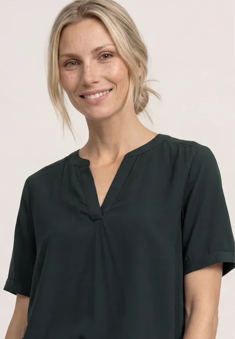 Een persoon draagt een donkergroene blouse met korte mouwen en V-hals tegen een lichte achtergrond.