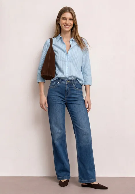 Frau in hellblauem Hemd, blauen Jeans, brauner Tasche und braunen Schuhen vor neutralem Hintergrund.