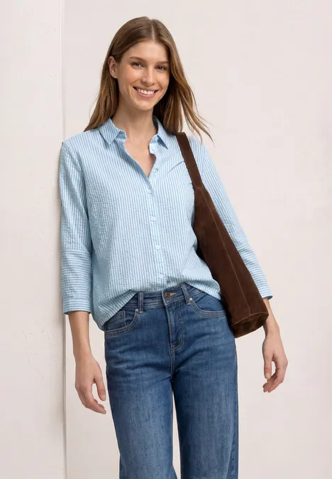 Frau mit hellblauem Hemd, dreiviertel Ärmeln, blauen Jeans und brauner Umhängetasche.