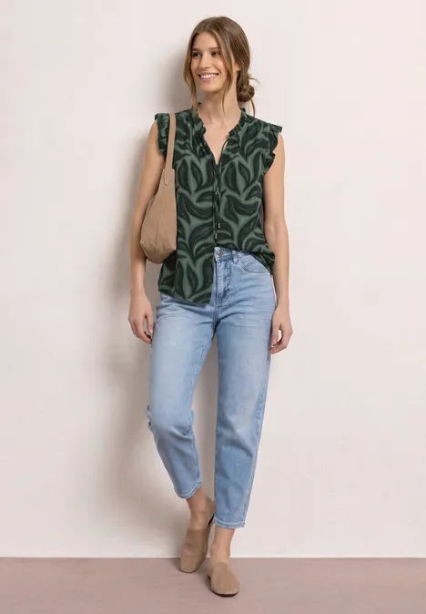 Frau in grün gemusterter Bluse, hellen Jeans und braunen Slippern mit Umhängetasche vor neutralem Hintergrund.