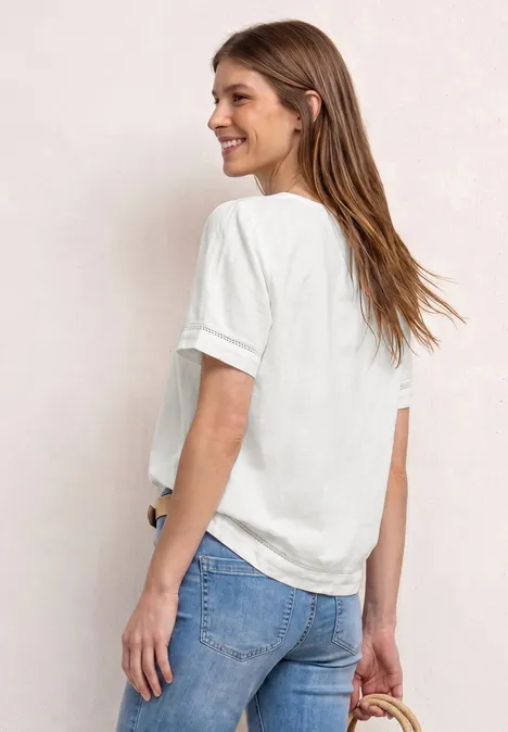 Eine Frau von hinten in weißem T-Shirt und blauen Jeans vor hellem Hintergrund.