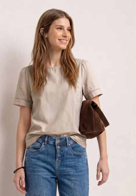 Eine Person in beige T-Shirt und Jeans, mit einer braunen Tasche über dem Arm, vor neutralem Hintergrund.