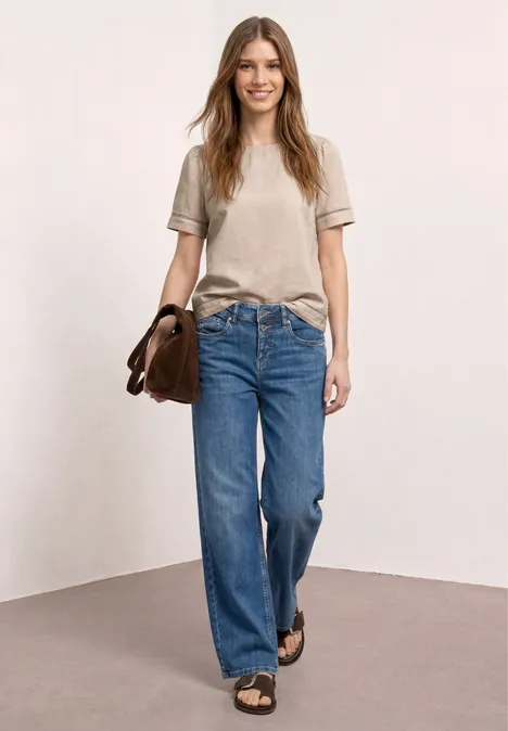 Frau in beiger T-Shirt, weiten Jeans und Sandalen, mit brauner Tasche vor neutralem Hintergrund.