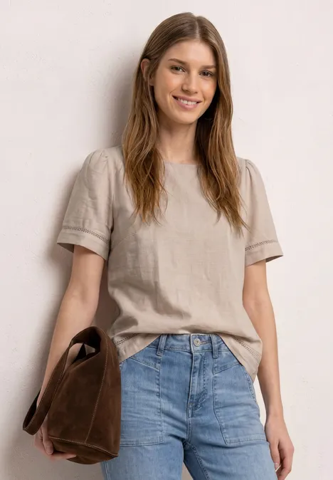 Frau steht in beigem T-Shirt und Jeans, hält eine braune Tasche.