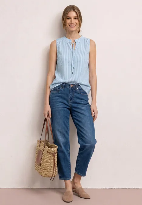 Frau trägt hellblaue ärmellose Bluse, blaue Jeans und Basttasche vor neutralem Hintergrund.