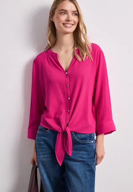 Frau mit pinker Bluse, vorne geknotet, und blauen Jeans vor hellem Hintergrund.
