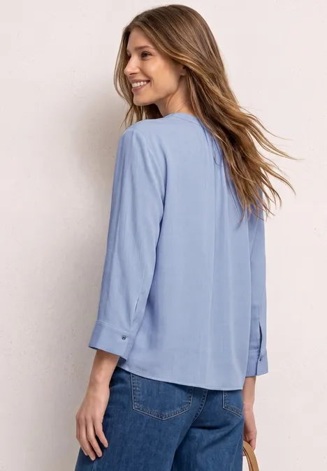 Vrouw van achter in lichtblauw blouse met 3/4 mouwen en jeans, lichte achtergrond.